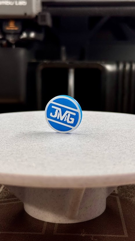Round blue and white JMG logo pin displayed on a gray pedestal.