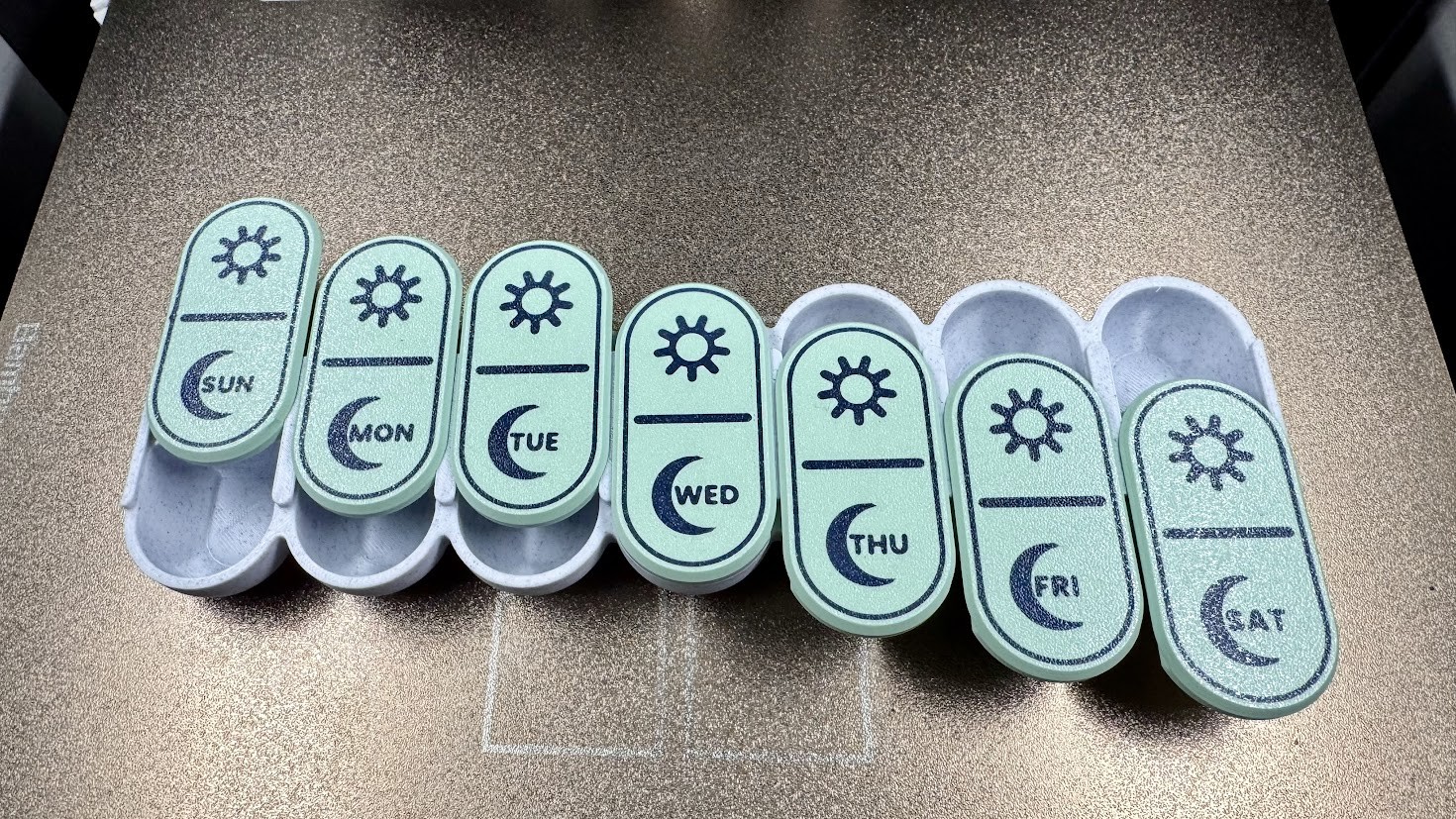 Weekly pill container with seven pale green lids labeled Sunday through Saturday, gray compartments, and sun and moon symbols.